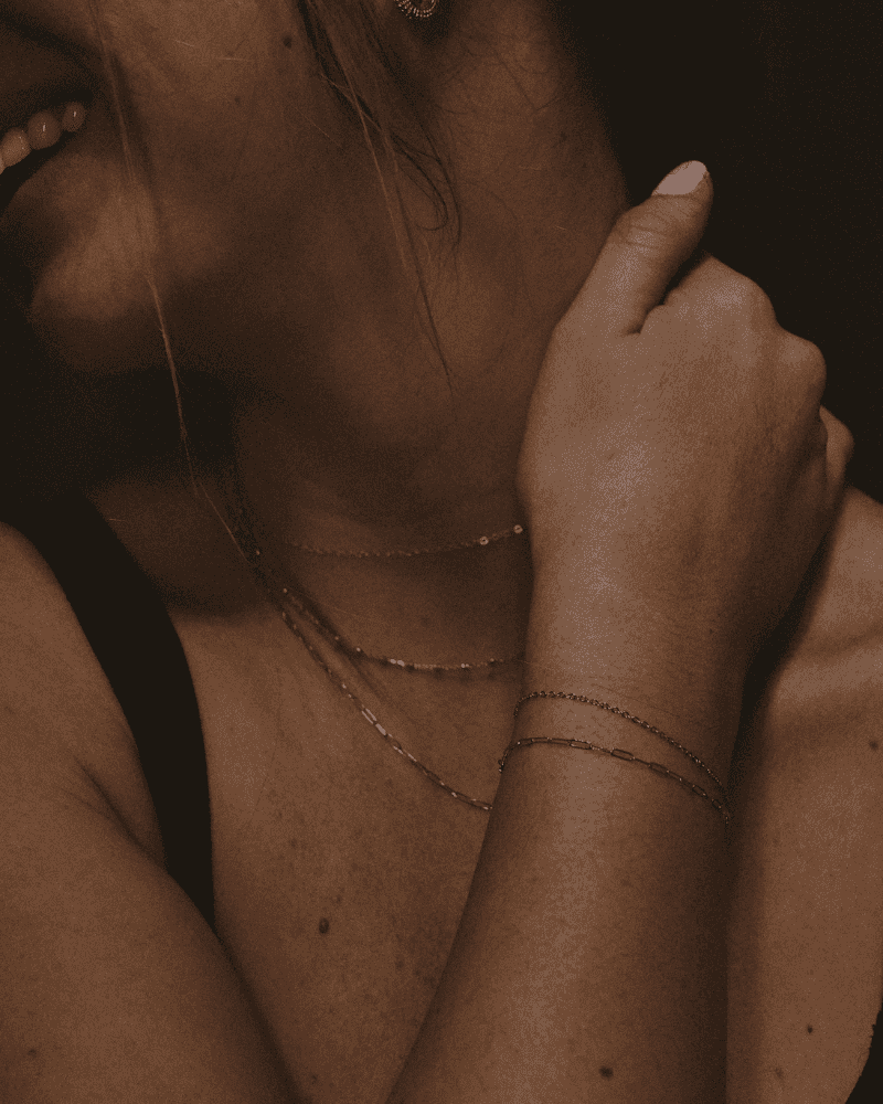 Close-up of a woman touching her neck and shoulder wearing thin necklaces and bracelets.